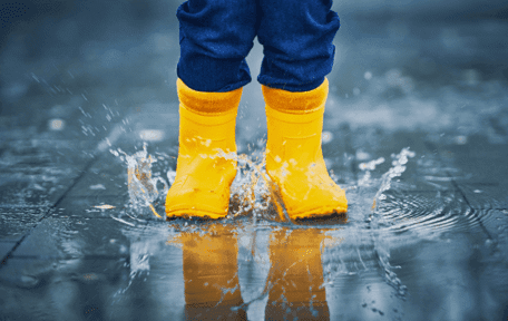 Kid in yellow rain boots walking on a lightly flooded walkway.
